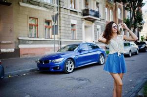 ropa de niña elegante y rizada en falda de jeans azules, blusa y gafas posadas cerca de un auto azul en la calle de la ciudad. foto