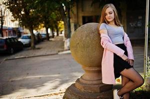 Young blonde girl in black skirt and pink coat posed at sunny day on street. photo