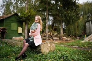 Young blonde girl at pink coat posed on autumn park, sitting on cut tree against house of the forester. photo