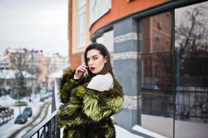 chica morena con abrigo de piel verde en la calle de la ciudad contra la casa con grandes ventanales en invierno. foto