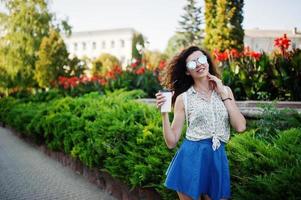 ropa de niña con estilo rizado en falda de jeans azules, blusa y gafas posadas en la calle de la ciudad con una taza de café. foto