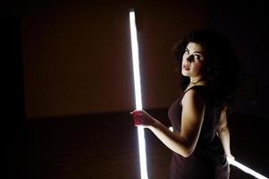 Curly brunette plus size model with two long led lamp at black room. photo