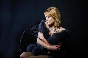 Studio portrait of sexy blonde girl with originally make up on neck and tattoo on thigh, wear on luxurious fur coat at dark background, sitting on chair.