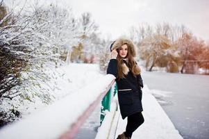 hermosa chica morena en ropa de abrigo de invierno. modelo en chaqueta de invierno contra el lago congelado en el parque. foto