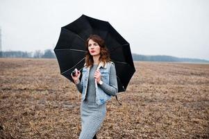 Portrait of brunette curly girl in jeans jacket with black umbrella at field. photo