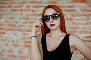 chica elegante de pelo rojo con gafas de sol vestidas de negro, contra un lugar abandonado con paredes de ladrillo. foto
