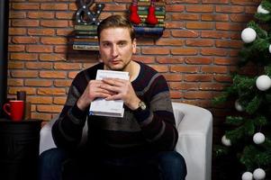 Studio portrait of man with book sitting on chair against christmass tree with decorations. photo