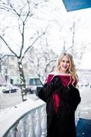 chica rubia elegante en vestido de noche rojo y abrigo de piel en las calles de la ciudad en el día de invierno. foto