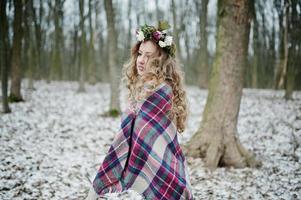 Curly cute blonde girl with wreath in checkered plaid at snowy forest in winter day. photo