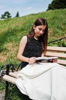 Portrait of a fabulous woman in black polka dot dress sitting on the bench with a huge white shawl and reading in the park. photo