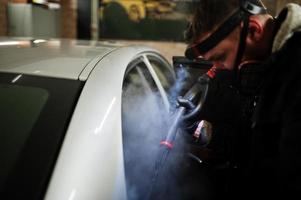 Man in uniform and respirator, worker of car wash center, cleaning car interior with hot steam cleaner. Car detailing concept. photo