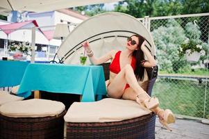 retrato de una chica impresionante en traje de baño de bikini rojo y gafas de sol disfrutando de su tiempo sentada en el sofá en el muelle junto al lago. foto