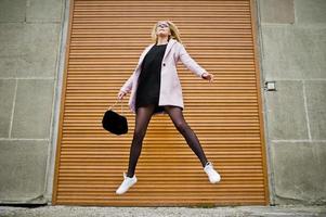 Blonde girl at glasses and pink coat with handbag jump against shutters. photo