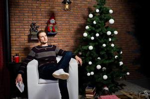 retrato de estudio de un hombre con un libro sentado en una silla contra un árbol de navidad con adornos. foto