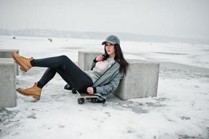chica morena elegante con gorra gris, estilo casual de calle con patineta en el día de invierno. foto