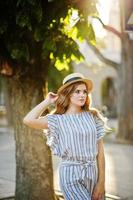 Portrait of a very attractive young woman in striped overall posing with her hat on a pavement in a town with trees in a background. photo