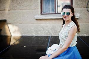 ropa de niña con estilo rizado en falda de jeans azules, blusa y gafas de sol. retrato en las calles de la ciudad. foto