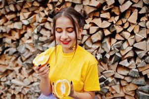 Young funny girl with bright make-up, wear on yellow shirt hold picece of orange against wooden background. photo