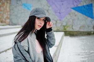 Stylish brunette girl in gray cap, casual street style on winter day against colored wall. photo