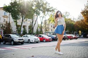 Curly stylish girl wear on blue jeans skirt, blouse and glasses posed at street of city with cup of coffee. photo