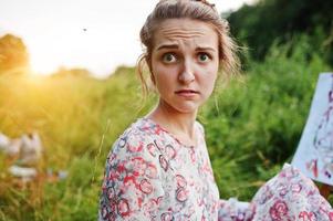 Portrait of a funny girl making weird facial expressions while painting on the meadow. photo