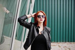 Red haired stylish girl in sunglasses wear in black, against large windows. Street fashion portrait.