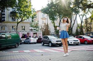 Curly stylish girl wear on blue jeans skirt, blouse and glasses posed at street of city with cup of coffee. photo