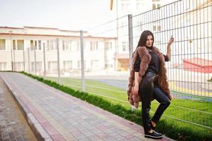 Fashion outdoor photo of gorgeous sensual woman with dark hair in elegant clothes and luxurious sleeveless fur coat against iron fence at autumn city.