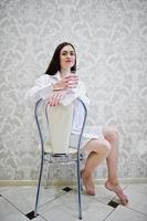 Portrait of a pretty young woman in male shirt and underwearsitting with a glass of water in her hands. photo