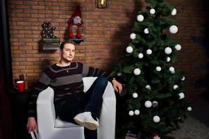 retrato de estudio de un hombre con un libro sentado en una silla contra un árbol de navidad con adornos. foto