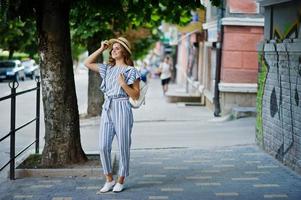 Portrait of a beautiful model in striped overall posing with hat and a backpack on a street with trees in a town. photo