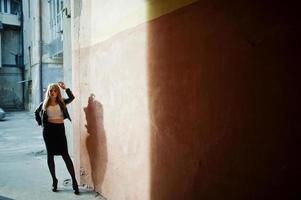 elegante chica rubia vestida con una chaqueta de cuero negro posando en las calles de la antigua muralla de fondo de la ciudad con sombras. foto