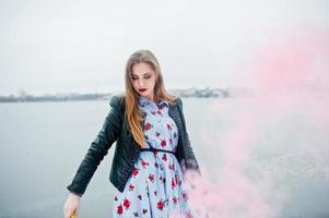 Stylish girl in leather jacket hold pink smoke flare at winter day against frozen lake. photo