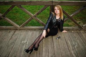 Elegant blonde girl wear on black leather jacket posing at streets of town sitting on wooden. photo