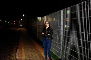 Night portrait of girl model wear on jeans and leather jacket against iron fence. photo