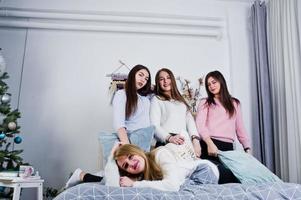 Four cute friends girls wear on warm sweaters and black pants on bed at new year decorated room on studio, play with pillows. photo