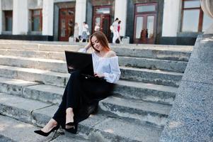 Good-looking young woman in white blouse, wide black pants and black classic high heels sitting on stairs and working on her laptop. photo