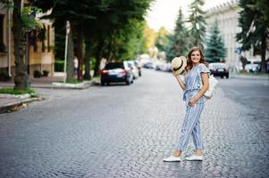 Portrait of a beautiful model in striped overall posing with hat and a backpack on a street with trees in a town. photo