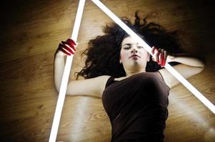 Curly brunette plus size model with two long led lamp at black room lying on flat. photo