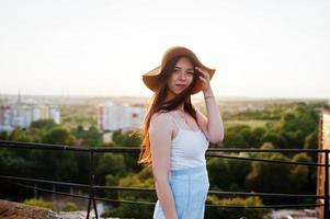 retrato de una mujer joven y bonita con camiseta blanca y falda azul posando en la azotea con su sombrero naranja al atardecer. foto