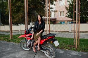 retrato de una mujer genial e impresionante con vestido y chaqueta de cuero negro sentada en una moto roja genial. foto