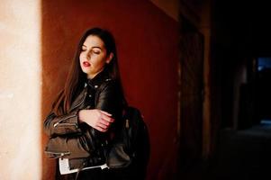 Stylish brunette girl wear on leather jacket and shorts with backpack against orange wall at shadows. photo