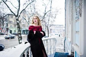chica rubia elegante en vestido de noche rojo y abrigo de piel en las calles de la ciudad en el día de invierno. foto