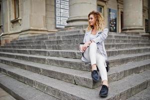 elegante modelo rubia rizada vestida de blanco con una taza de café posando en las escaleras al aire libre. foto