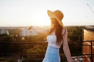 retrato de una joven impecable con camiseta blanca, falda azul y sombrero naranja apuntando al sol con la mano mientras está de pie en la azotea. foto