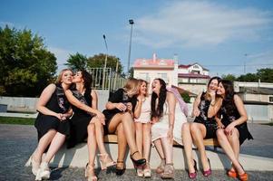 Group of 7 girls wear on black and 2 brides sitting on bench and having fun at hen party. photo