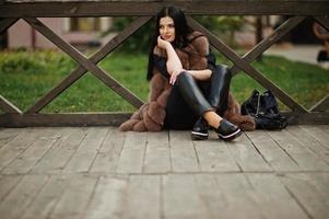 Fashion outdoor photo of gorgeous sensual woman with dark hair in elegant clothes and luxurious fur coat sitting against wooden railings.