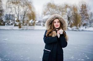 Beautiful brunette girl in winter warm clothing. Model on winter jacket against frozen lake at park. photo