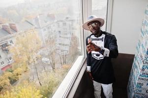 Rich african man looking at panoramic window at his penthouse. Portrait of successful black man in hat indoor. photo