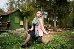 Young blonde girl at pink coat posed on autumn park, sitting on cut tree against house of the forester. photo